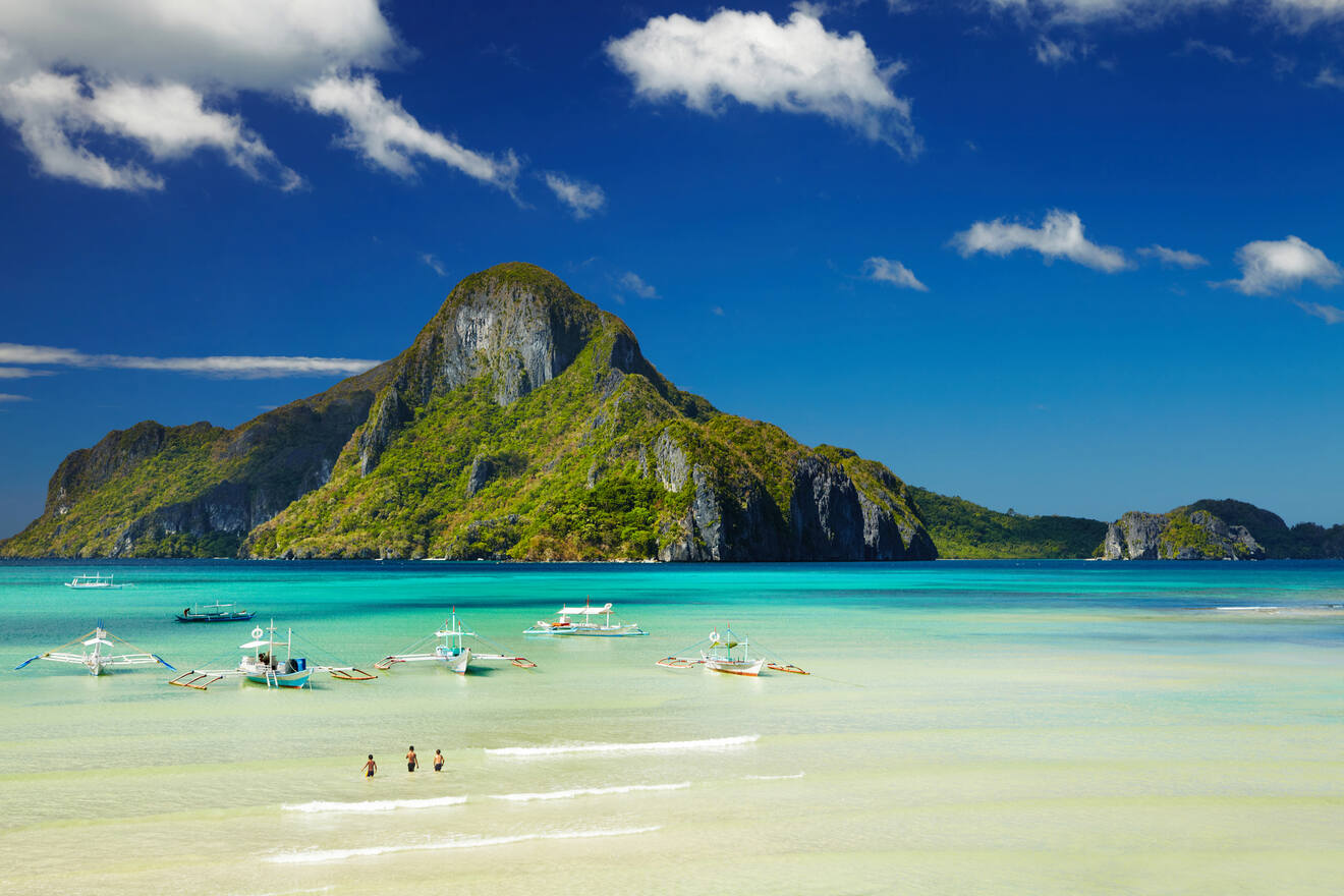 Turquoise waters with traditional Filipino boats anchored near a sandy beach under a clear blue sky, overlooked by a majestic limestone mountain.